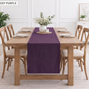 Op de afbeelding: Een dieppaarse rechthoekige tafelloper in het midden van een lichtbruine houten eettafel. De loper is gemaakt van een effen gekleurde stof. De tafel is gedekt met vier witte borden en in het midden staat een witte vaas met bloemen.