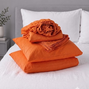 May include: A stack of bright orange bedding consisting of two square pillowcases and a fitted sheet with elasticized edges. The fabric has a visible woven texture. The text BRIGHT ORANGE is printed in a white box in the upper left corner.