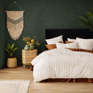 May include: A bedroom scene featuring a bed with white bedding and a textured, arched pattern. Brown accent pillows and a brown bed skirt complement the white duvet. A woven macrame wall hanging and a rattan nightstand with a vase of flowers add to the decor.