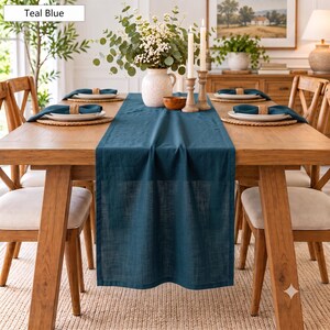 May include: A teal blue linen table runner draped over a wooden dining table. The table is set with woven placemats, white plates, and dark teal napkins. A white ceramic vase with eucalyptus and white flowers sits in the centre.