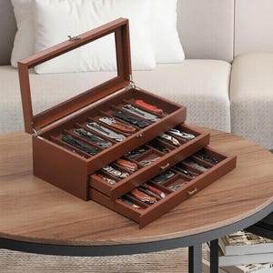 May include: A wooden knife display case with a glass top, showcasing multiple pocket knives. The case is brown with three pull-out drawers and individual compartments for each knife. The knives have various handle colors and blade designs.