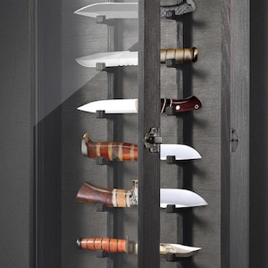 May include: A dark gray wooden display case with a glass door, showcasing an assortment of knives. The knives have various blade shapes and handle designs, including wood and metal. The case is mounted on a dark gray wall.