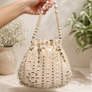 May include: A beige drawstring handbag featuring intricate silver-toned mirror work in geometric patterns across the exterior. The bag includes a handle made of large, round, cream-colored beads and a bottom edge finished with small, dangling silver accents.