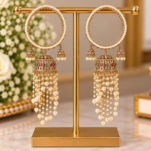 May include: A pair of gold-toned hoop earrings featuring a circular frame lined with small white pearls. Each earring includes a central bell-shaped pendant adorned with red and green stones, accented by dangling pearl strands and small bells.