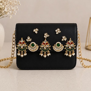 May include: A black rectangular shoulder bag featuring a gold-tone chain strap. The front flap is decorated with five ornate gold-tone metal embellishments, each set with green, red, and white stones, accented with small white faux pearls.