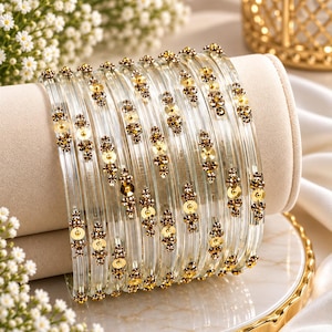 May include: A set of clear glass bangles featuring gold-toned metallic accents and small brown crystals arranged in a repeating floral pattern. The circular bracelets are arranged in a stack, showcasing the transparent material and the decorative embellishments.