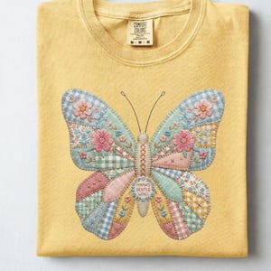 Può includere: Una maglietta gialla con un disegno a farfalla. La farfalla è realizzata in tessuto patchwork dai colori pastello, con motivi floreali e a quadretti. Il testo "Mama's Gentle Wing" è sulla farfalla. La parola "Butter" è in basso.