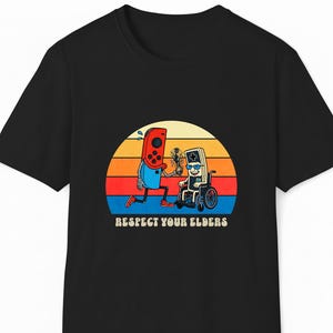 Op de afbeelding: Zwart T-shirt met een retro afbeelding van twee gamecontrollers. Eén controller rent en de andere zit in een rolstoel. De tekst "Respect Your Elders" staat eronder, tegen een zonsondergang.
