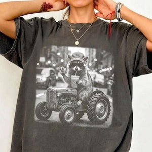 以下が含まれることがあります： チャコールグレーの半袖Tシャツ。野球帽とサングラスを着用し、トラクターに乗って飲み物を持っているアライグマの白黒グラフィックプリントが施されています。クルーネックでゆったりとしたフィット感のTシャツです。