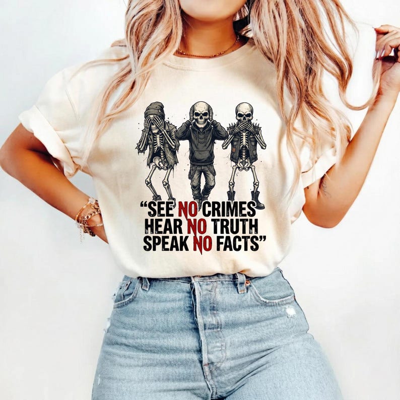 May include: Cream-colored t-shirt featuring a graphic of three skeletons in the 'see no evil, hear no evil, speak no evil' pose. The text "SEE NO CRIMES HEAR NO TRUTH SPEAK NO FACTS" is printed below the skeletons in black and red.