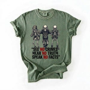 May include: Green t-shirt featuring a graphic of three skeletons in various poses, with one covering its eyes, one its ears, and one its mouth. The text "SEE NO CRIMES HEAR NO TRUTH SPEAK NO FACTS" is below the skeletons.