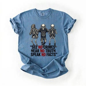 May include: A light blue t-shirt featuring a graphic of three skeletons in the 'see no evil, hear no evil, speak no evil' pose. The text on the shirt reads, "SEE NO CRIMES, HEAR NO TRUTH, SPEAK NO FACTS."
