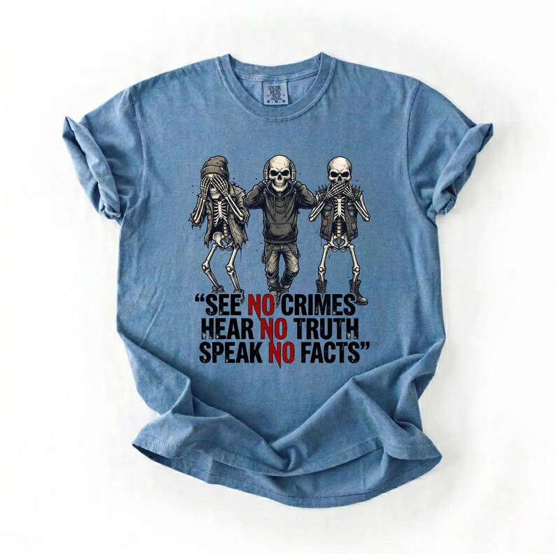 May include: A light blue t-shirt featuring a graphic of three skeletons in the 'see no evil, hear no evil, speak no evil' pose. The text on the shirt reads, "SEE NO CRIMES, HEAR NO TRUTH, SPEAK NO FACTS."