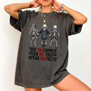 May include: Dark gray t-shirt featuring a graphic of three skeletons in the 'see no evil, hear no evil, speak no evil' pose. The text below reads "SEE NO CRIMES, HEAR NO TRUTH, SPEAK NO FACTS".