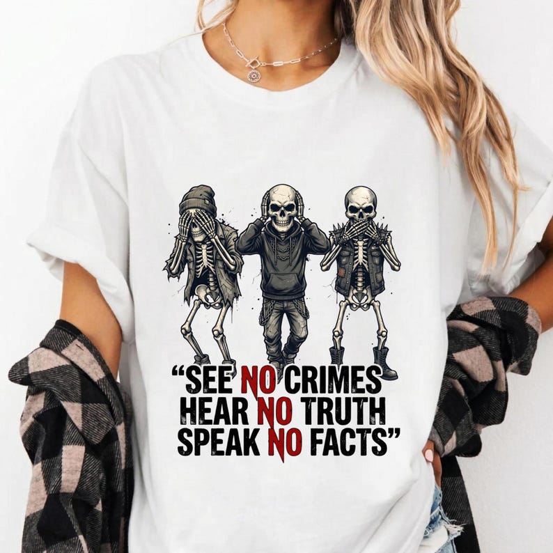 May include: White t-shirt featuring a graphic of three skeletons in various poses, with the text "SEE NO CRIMES HEAR NO TRUTH SPEAK NO FACTS" in black and red. The skeletons are in a cartoon style.