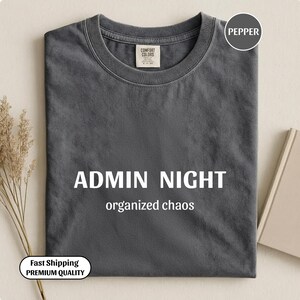 Può includere: Una maglietta grigio scuro con le parole "ADMIN NIGHT" e "organized chaos" stampate in bianco. La maglietta ha un girocollo e un'etichetta sul colletto.