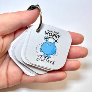 Puede incluir: Un conjunto de tarjetas cuadradas blancas en un anillo de metal. La tarjeta superior presenta un monstruo de dibujos animados azul con dos ojos y el texto "MANAGING WORRY WITH Jitters" impreso en negro. Las tarjetas tienen esquinas redondeadas.