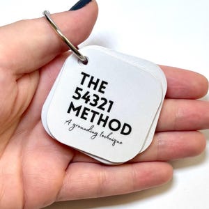 Puede incluir: Un conjunto de tarjetas cuadradas blancas con esquinas redondeadas unidas por una anilla de metal plateado. La tarjeta superior presenta texto negro que dice THE 54321 METHOD y A grounding technique en una fuente cursiva.