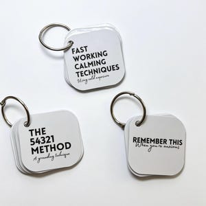 Könnte beinhalten: Drei Sätze weißer quadratischer Karten mit abgerundeten Ecken, die von silbernen Metallringen zusammengehalten werden. Die Karten tragen schwarzen Text: FAST WORKING CALMING TECHNIQUES Using cold exposure, THE 54321 METHOD A grounding technique und REMEMBER THIS When you're anxious.