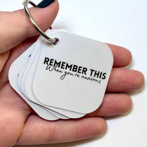 May include: A set of white square cards with rounded corners, held together by a silver metal ring. The top card features the black printed text REMEMBER THIS When you're anxious. The cards are made of a smooth, matte material and are stacked.
