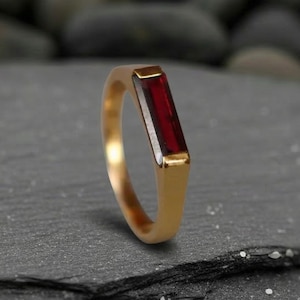 Może przedstawiać: Złoty pierścionek z prostokątnym, głęboko czerwonym kamieniem szlachetnym. Pierścionek ma prosty, nowoczesny design z polerowanym wykończeniem. Kamień jest osadzony równo z obrączką. Pierścionek jest prezentowany na ciemnoszarej powierzchni.