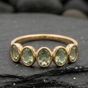 May include: A gold ring featuring six oval-cut, light green gemstones. The ring's band is a thin, polished gold. The gemstones are set in a row, creating a delicate and elegant design. The ring is displayed on a dark, textured surface.