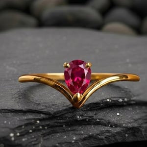 Könnte beinhalten: Ein goldener Ring mit einem birnenförmigen, tiefroten Edelstein. Das Band des Rings bildet ein V, wobei der Edelstein an der Spitze sitzt. Der Ring wird auf einer dunklen, strukturierten Oberfläche präsentiert.