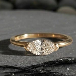 Op de afbeelding: Een gouden ring met een marquise-geslepen diamant in een bezel-zetting. De ring ligt op een donkergrijze ondergrond met een vage achtergrond van donkergrijze stenen. De diamant is helder en sprankelend.