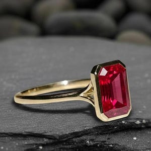 May include: A gold ring featuring a large, rectangular, red gemstone. The gemstone is set in a bezel setting, and the band has a simple, elegant design. The ring is displayed on a dark, textured surface.