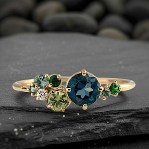 Può includere: Un anello d'oro con una grande gemma rotonda blu scuro come punto focale, circondata da gemme verdi e bianche più piccole. La fascia dell'anello è in oro lucido e sottile, e le gemme sono incastonate in un design a grappolo.