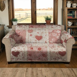 May include: A floral patterned sofa cover with a patchwork design. The cover features a repeating pattern of pink hearts and roses on a cream background. The sofa is a neutral beige colour.