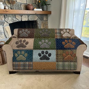 May include: A beige sofa with a paw print patterned cover. The cover is made of patchwork squares in various colors, including brown, green, and blue. Each square features a large paw print design, creating a cozy and pet-friendly aesthetic.