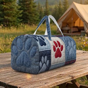 Könnte beinhalten: Eine gesteppte Denim-Duffel-Tasche mit einem Patchwork-Design mit großen blauen und roten Pfotenabdrücken. Die Tasche hat weiße Griffe und steht auf einem Picknicktisch aus Holz. Im Hintergrund sind ein Zelt und Bäume zu sehen.
