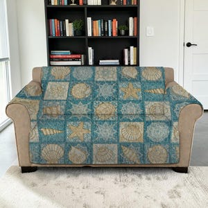 May include: A blue and beige sofa cover featuring a repeating pattern of seashells, starfish, and geometric designs. The cover is draped over a beige sofa, creating a coastal theme. The background includes a bookshelf and a white door.