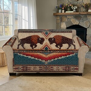 May include: A beige sofa with a decorative cover featuring a Native American design. The cover displays two brown bison facing each other, set against a backdrop of geometric patterns in teal, red, and beige. The sofa is in a living room setting.