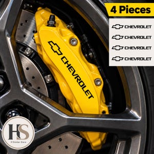 Op de afbeelding: Een set van vier Chevrolet-stickers met zwarte tekst en het vlinderdaslogo, ontworpen voor toepassing op remklauwen. De stickers worden getoond naast een felgele remklauw gemonteerd op een autowiel met een donkere metalen velg.