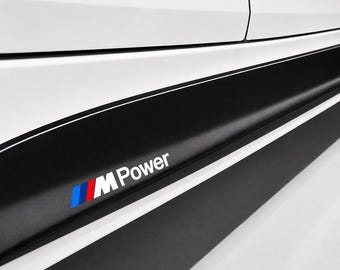 Puede incluir: Un panel lateral de coche blanco con una calcomanía horizontal negra en el faldón lateral y una fina línea blanca. La calcomanía muestra el logotipo M Power en texto blanco, junto a un gráfico de rayas inclinadas en azul, morado y rojo.