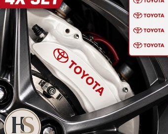 Puede incluir: Un juego de cuatro pegatinas blancas para pinzas de freno Toyota con letras rojas y el logotipo de Toyota. Las pegatinas se muestran aplicadas sobre una pinza de freno blanca detrás de una rueda de coche gris oscuro. Texto 4X SET y cuatro logos.
