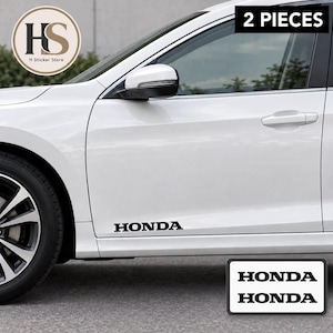 May include: A set of two black vinyl decals featuring the word HONDA in a bold, capitalised font. The stickers are designed for application on the lower side panel of a white car, as shown in the image, and include the text 2 PIECES.