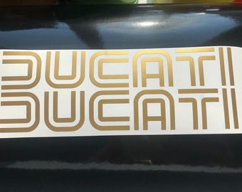 Puede incluir: Calcomanías "DUCATI" doradas sobre fondo blanco. La palabra se repite dos veces, con una fuente retro y geométrica. Las calcomanías son probablemente para una motocicleta o vehículo.