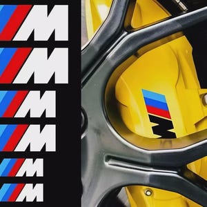 Op de afbeelding: Afbeelding toont meerdere witte "M" logo's met blauwe, rode en witte strepen, en een gele remklauw met een bijpassend logo. De logo's zijn waarschijnlijk voor een BMW-voertuig, wat duidt op auto-accessoires.