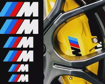 Puede incluir: La imagen presenta múltiples logotipos "M" blancos con rayas azules, rojas y blancas, y una pinza de freno amarilla con un logotipo a juego. Los logotipos son probablemente para un vehículo BMW, lo que sugiere accesorios automotrices.