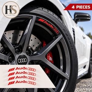 May include: A set of four red Audi rim decals featuring the brand logo and four rings, designed for application on car wheels. The stickers are shown applied to a dark grey alloy wheel with a red brake caliper and a black tyre on a white car.