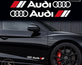 Puede incluir: Dos calcomanías blancas de Audi con el nombre de la marca y el logotipo de los cuatro aros junto a un gráfico de rayas inclinadas rojas y blancas. El juego incluye dos pegatinas, aplicadas en el panel lateral inferior de un coche negro.