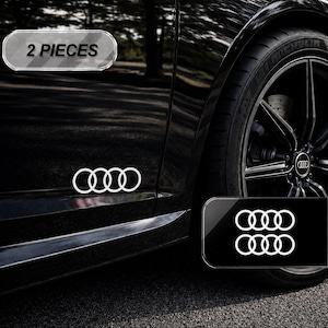 May include: A set of two white Audi four-ring logo decals applied to the side of a black car. The decals feature the iconic interlocking circle design. A rectangular overlay in the top left corner displays the text 2 PIECES in black lettering.