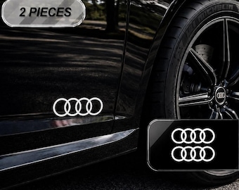 Puede incluir: Un juego de dos calcomanías blancas con el logotipo de los cuatro aros de Audi, aplicadas en el lateral de un coche negro. Las calcomanías presentan el diseño icónico de círculos entrelazados. Un recuadro muestra el texto 2 PIECES.