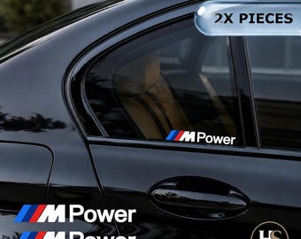 Puede incluir: Dos pegatinas de vinilo M Power con las icónicas franjas inclinadas en azul, morado y rojo seguidas del texto blanco M Power. Estos adhesivos están diseñados para su aplicación en ventanas o paneles de carrocería de un vehículo negro.