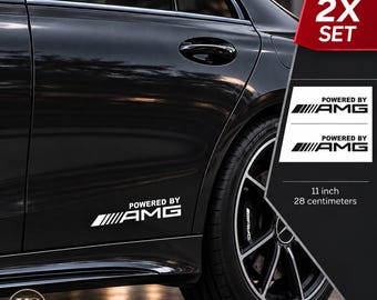 Puede incluir: Un juego de dos pegatinas de vinilo blanco para vehículo, cada una con el texto "POWERED BY AMG" y un elemento gráfico de rayas. Las pegatinas miden 28 centímetros de largo y se muestran aplicadas en el lateral de una puerta de coche negra.
