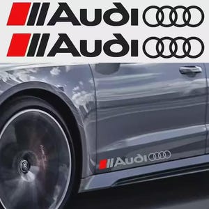 以下が含まれることがあります： 画像には、アウディの車のデカールが写っています。デカールには、黒い「Audi」という文字、赤い長方形、3本の黒い縦線が入ったアウディのロゴがあります。デカールはグレーの車に表示されています。