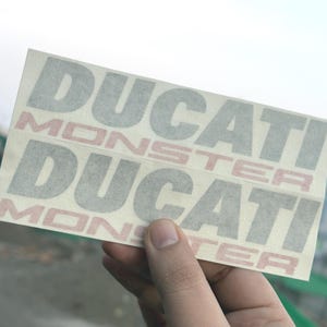 May include: Two Ducati Monster stickers are held up against a blurred outdoor background. The stickers are rectangular and feature the words "DUCATI" and "MONSTER" in a grey and red colour scheme. The stickers are held by a hand.
